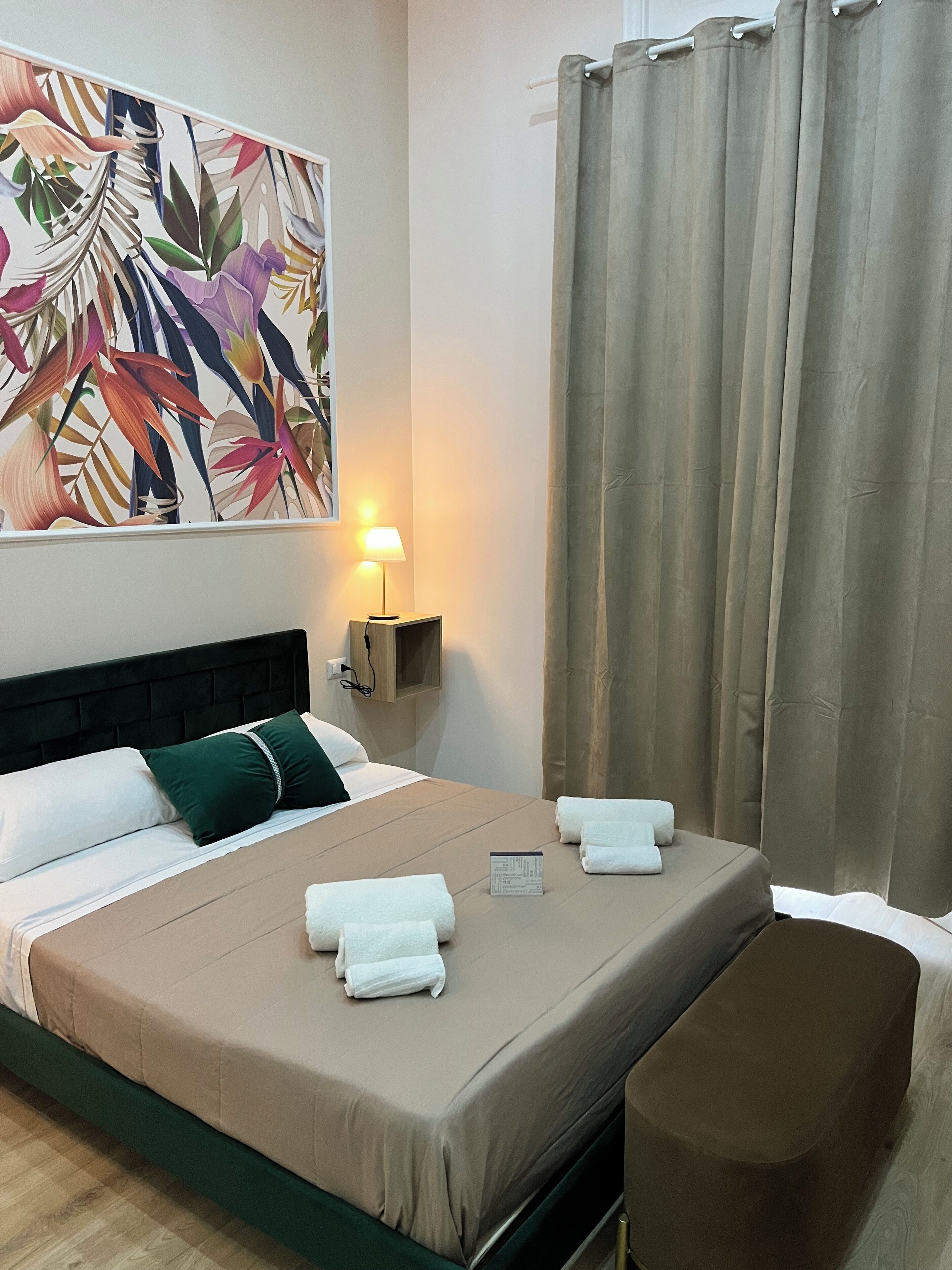 A hotel room with a bed, a nightstand with a lamp, a painting on the wall, and a window with curtains.
