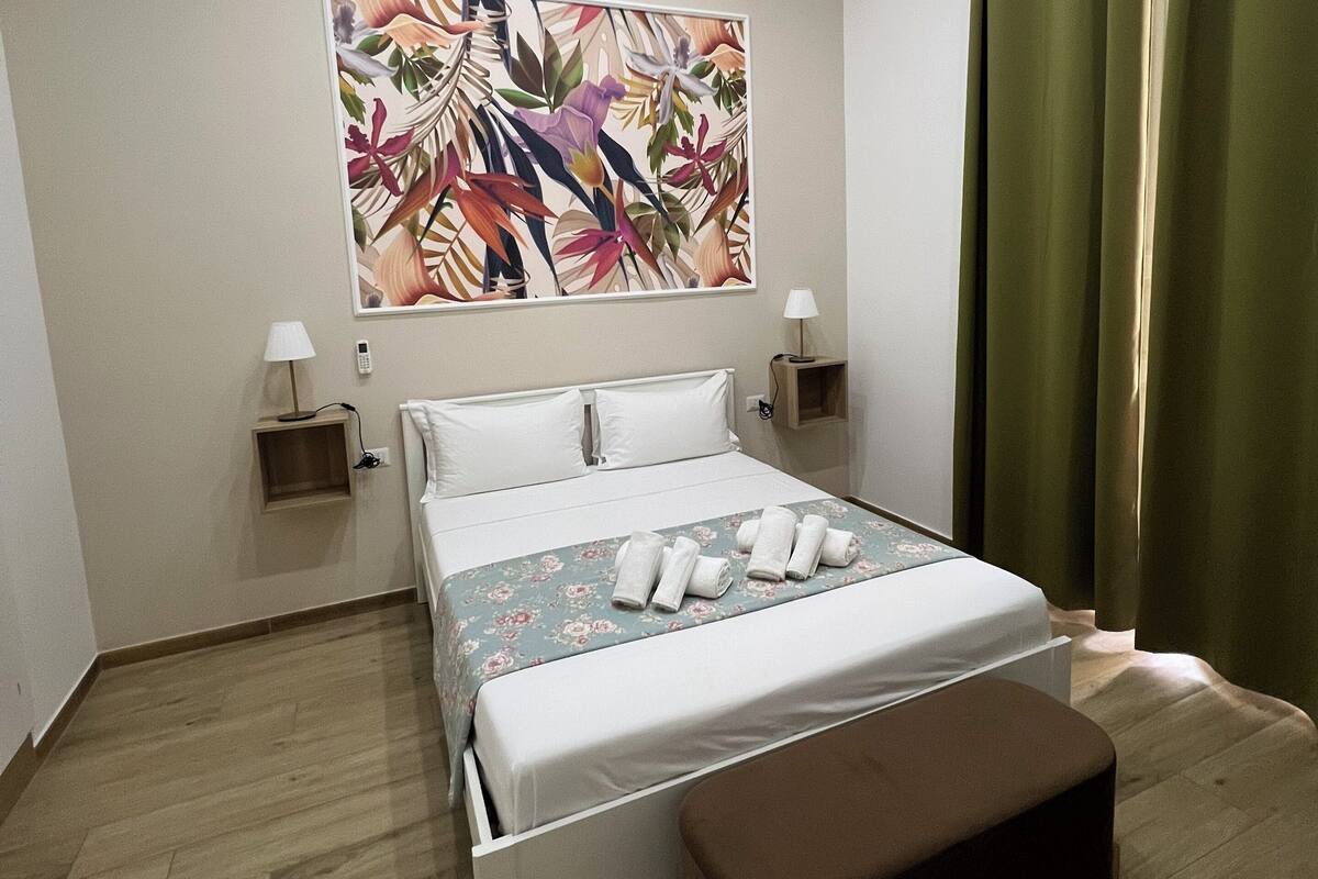 A modern hotel room with a bed, a bench, a nightstand, a lamp, and a large floral artwork on the wall.