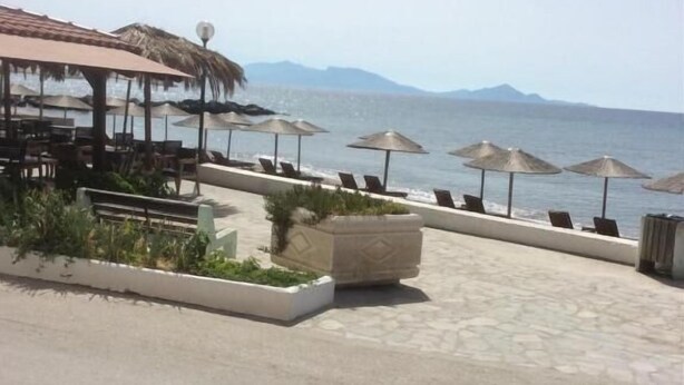 Un espace aménagé au bord de la plage, avec des sièges en plein air et des parasols, une structure à toit de chaume, et une vue sur la mer et les montagnes.