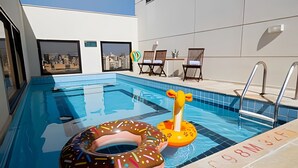 A swimming pool with two inflatable rings, one shaped like a donut and the other like a flamingo. There are lounge chairs and a view of a cityscape through the windows.