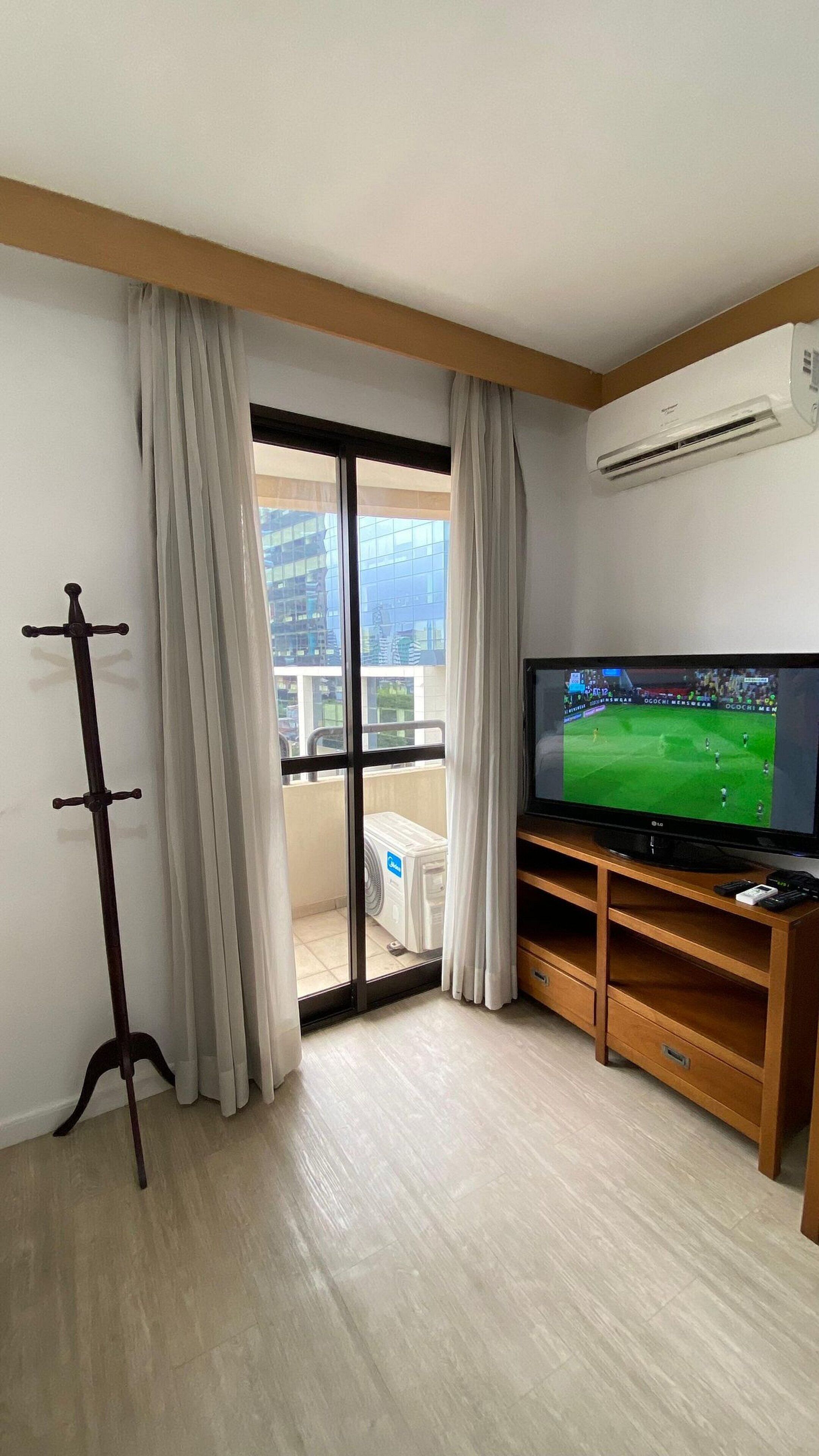 A living room with a TV, wooden entertainment unit, and a view of a cityscape through a large sliding glass door.