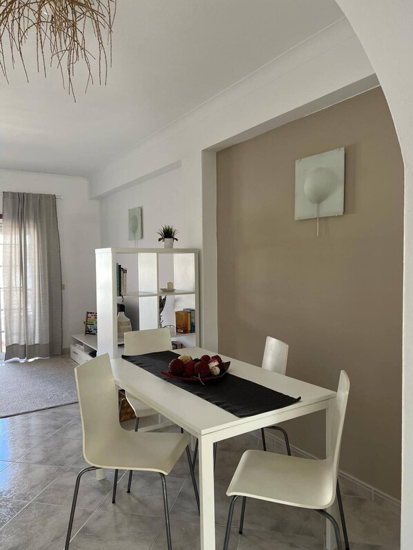A modern dining area with a white table and chairs, a decorative plant on a shelf, and a bookshelf in the background.