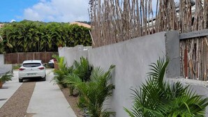 Un camino de concreto que lleva a un coche blanco estacionado en una entrada, bordeado por un muro con elementos de madera verticales y pequeñas plantas verdes.