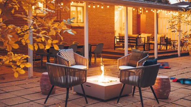 En udendørs patio med bålsted, fletstole og en murbygning med lyskæder.
