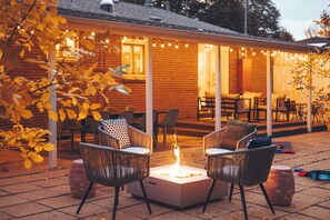 Een terras buiten met een vuurkorf, rieten stoelen en een bakstenen gebouw met lichtjes.