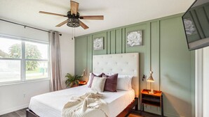 Una camera da letto con un letto, una ventola a soffitto, una finestra con tende e un comodino con una lampada.