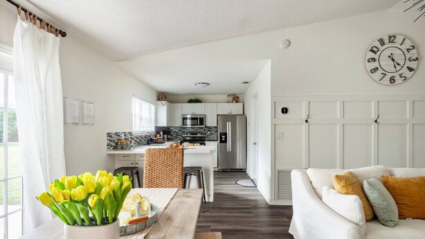 Una cucina moderna con mobili bianchi, un forno a microonde e un frigorifero. C'è una zona pranzo con un tavolo di legno e un vaso di tulipani gialli. Un divano bianco con cuscini colorati è in primo piano.