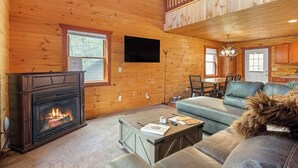 Ein gemütliches Blockhaus-Interieur mit Kamin, einem an der Wand befestigten Fernseher, einem Essbereich mit Tisch und Stühlen sowie einem Balkon mit Geländer.