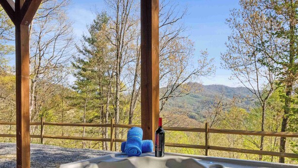 Ein Whirlpool mit einer Weinflasche und Handtüchern, umgeben von einer Holzterrasse und Bäumen, mit Blick auf eine gebirgige Landschaft.