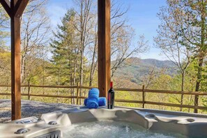 Un jacuzzi con una botella de vino y toallas, rodeado por una terraza de madera y árboles, con vistas a un paisaje montañoso.
