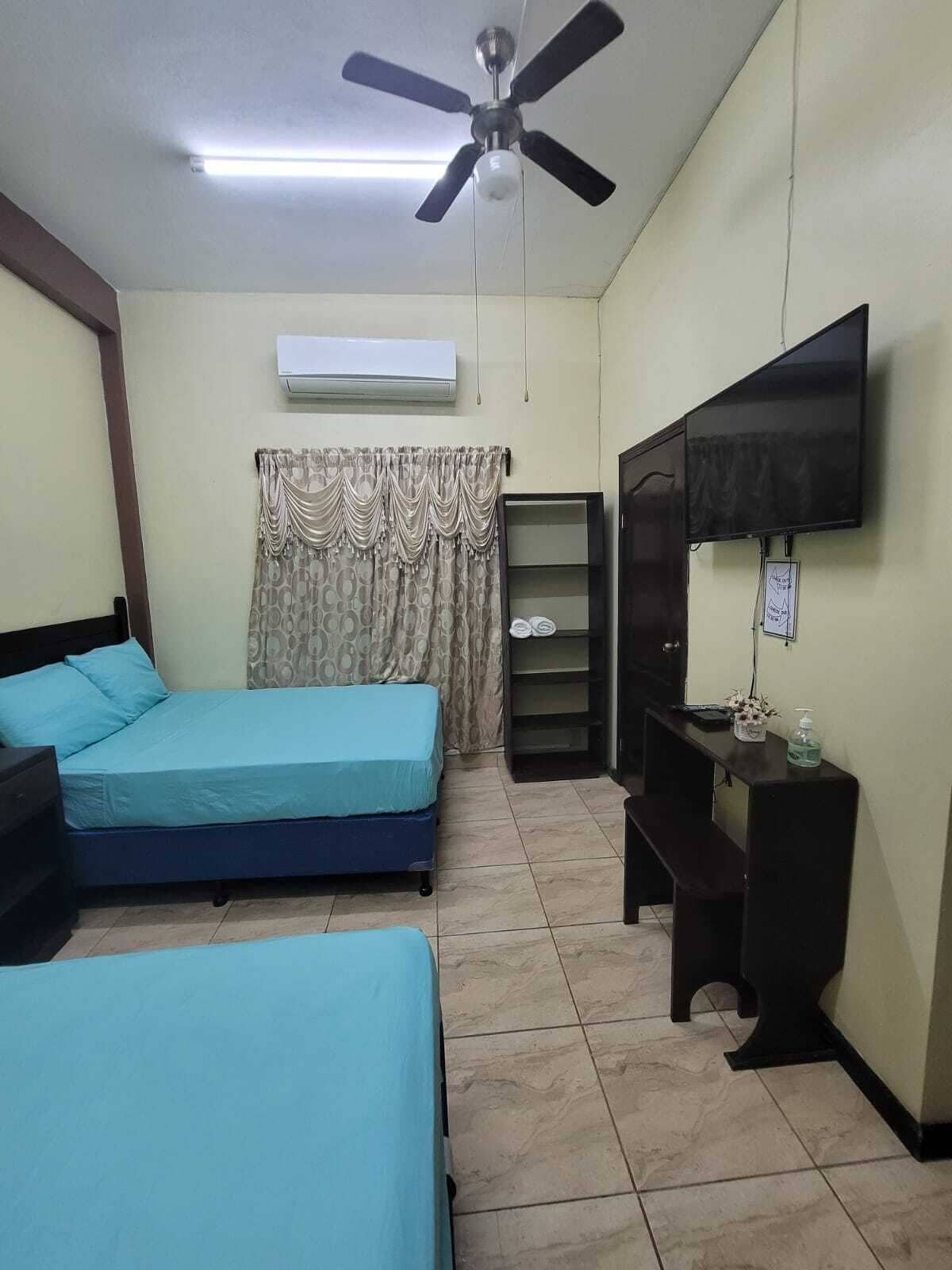 Een kamer met twee bedden, een plafondventilator, een airconditioning, een televisie en een klein tafeltje met handdesinfectiemiddel.