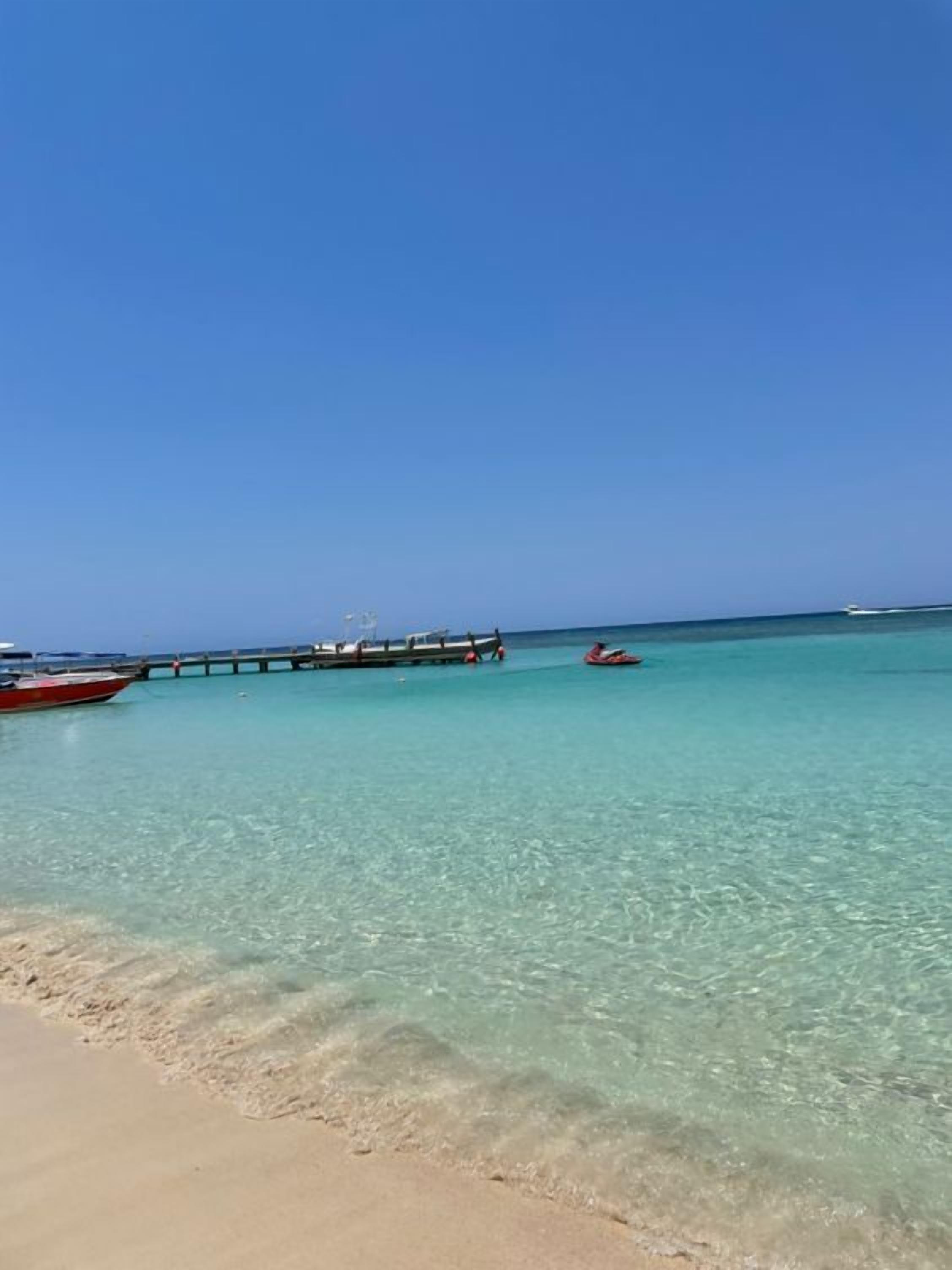 A beach with clear turquoise water, a pier, and boats.