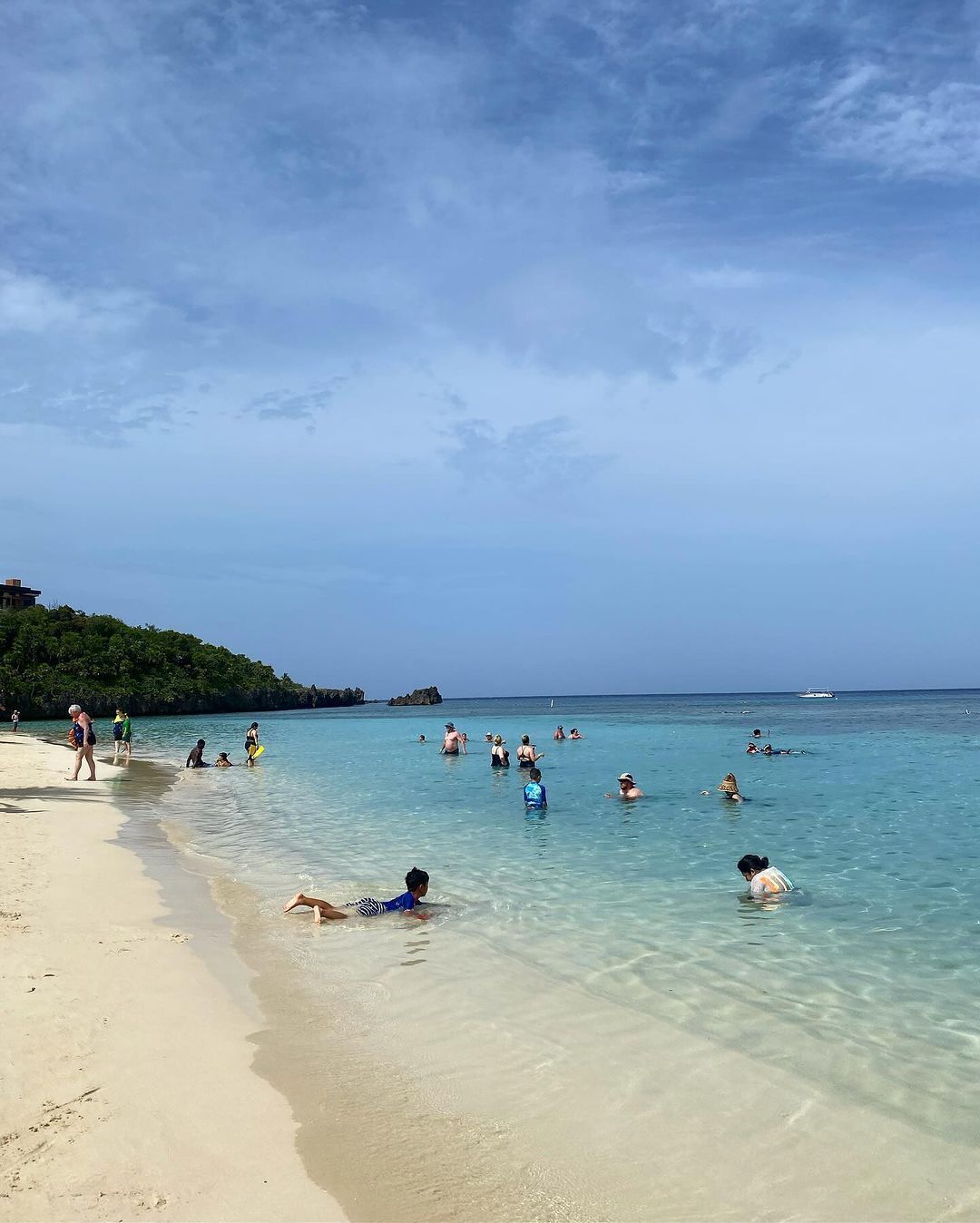 Una playa con agua cristalina, gente nadando y tomando el sol, y un barco a lo lejos.
