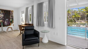 A modern living room with a black leather chair, a glass table, and a dining area with a wooden table and chairs. A large painting is on the wall, and there is a view of a pool outside. - Spacious Suite at New Yorker Hotel - Pool, Free Parking, WiFi and Coffee! (Miami)