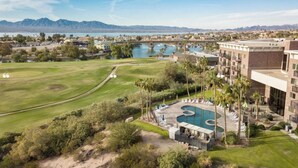 Campo de golf con zona de piscina, un puente sobre el agua y montañas al fondo.