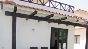 Une terrasse extérieure avec une table et des chaises en bois, une structure en forme de pergola et un bâtiment blanc avec un toit recouvert de tuiles.