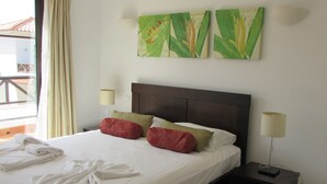 Un lit bien fait, avec du linge de lit blanc, des coussins rouges et verts, et un ensemble de serviettes blanches posées à côté. Deux lampes murales sont fixées de chaque côté du lit, et deux reproductions encadrées de motifs botaniques sont accrochées au-dessus.