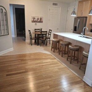 A kitchen with wooden flooring, a dining table with chairs, a bar area with stools, and a refrigerator.