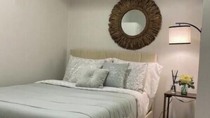 Un dormitorio con cama, un espejo redondo, una mesita de noche con lámpara y un pequeño florero con flores.