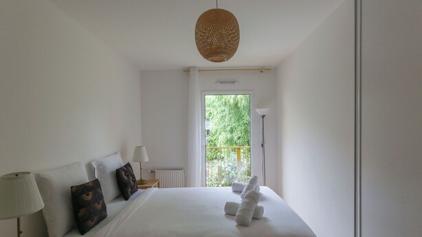 Une chambre avec un grand lit, une table de chevet avec une lampe, une fenêtre donnant sur un paysage verdoyant et un luminaire suspendu.