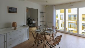 Un coin repas avec une table en verre, des chaises en osier et un buffet blanc. On aperçoit un balcon avec des balustrades jaunes.