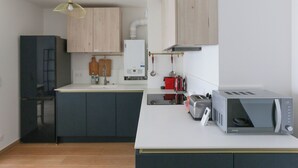 Une cuisine moderne avec un plan de travail blanc, un four à micro-ondes encastré, un grille-pain et une bouilloire. On trouve un meuble en bois au-dessus de l’évier et un luminaire suspendu.