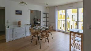 Un vaste espace dédié à la salle à manger, avec une table ronde et des chaises en osier, un buffet blanc avec des tiroirs, et une vue sur un bâtiment jaune, visible à travers de grandes fenêtres.