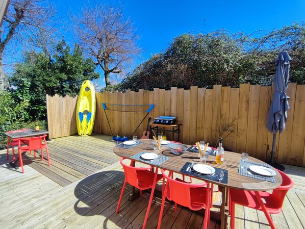 A wooden deck with a round table set for four, surrounded by red chairs. A kayak is leaning against the fence, and a barbecue grill is visible in the background.