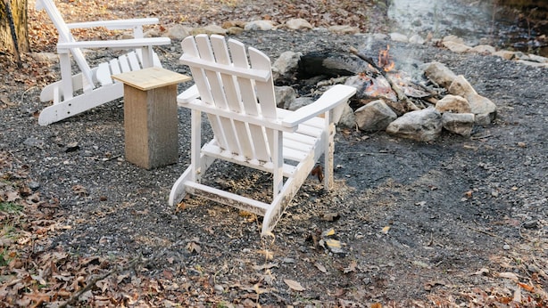 Een rustieke hut met een vuurkuil, twee Adirondack-stoelen en een kleine schuur, gelegen in een bebost gebied.