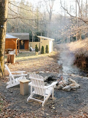 Eine rustikale Holzhütte mit Feuerstelle, zwei Adirondack-Stühlen und einem kleinen Schuppen in einem bewaldeten Gebiet.