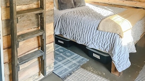 Een slaapkamer met een wand van houten latten, een bed met een grijze deken, een ladder en een ingelijst schilderij aan de muur.