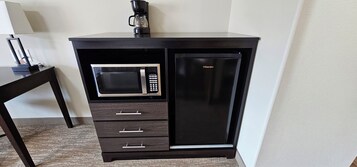 Un mini bar negro con un microondas y un refrigerador, una pequeña mesa con una lámpara, y una cafetera encima.