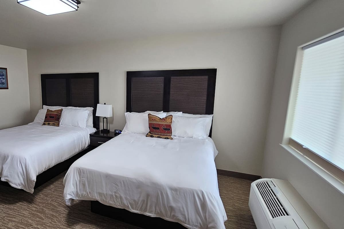 A hotel room with two beds, a wooden barn door, a nightstand with a lamp, and a window with blinds.
