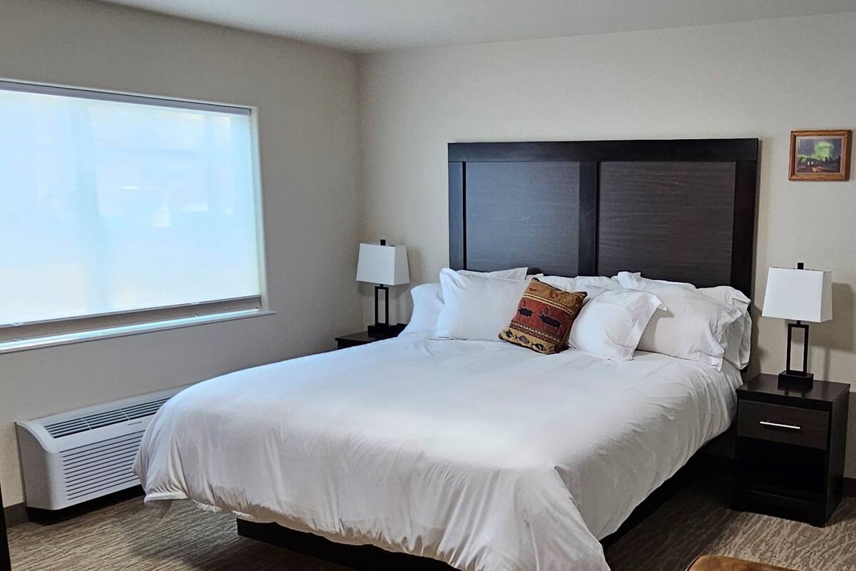 A neatly made bed with white linens, two bedside tables with lamps, a window with blinds, and a wall-mounted air conditioner.