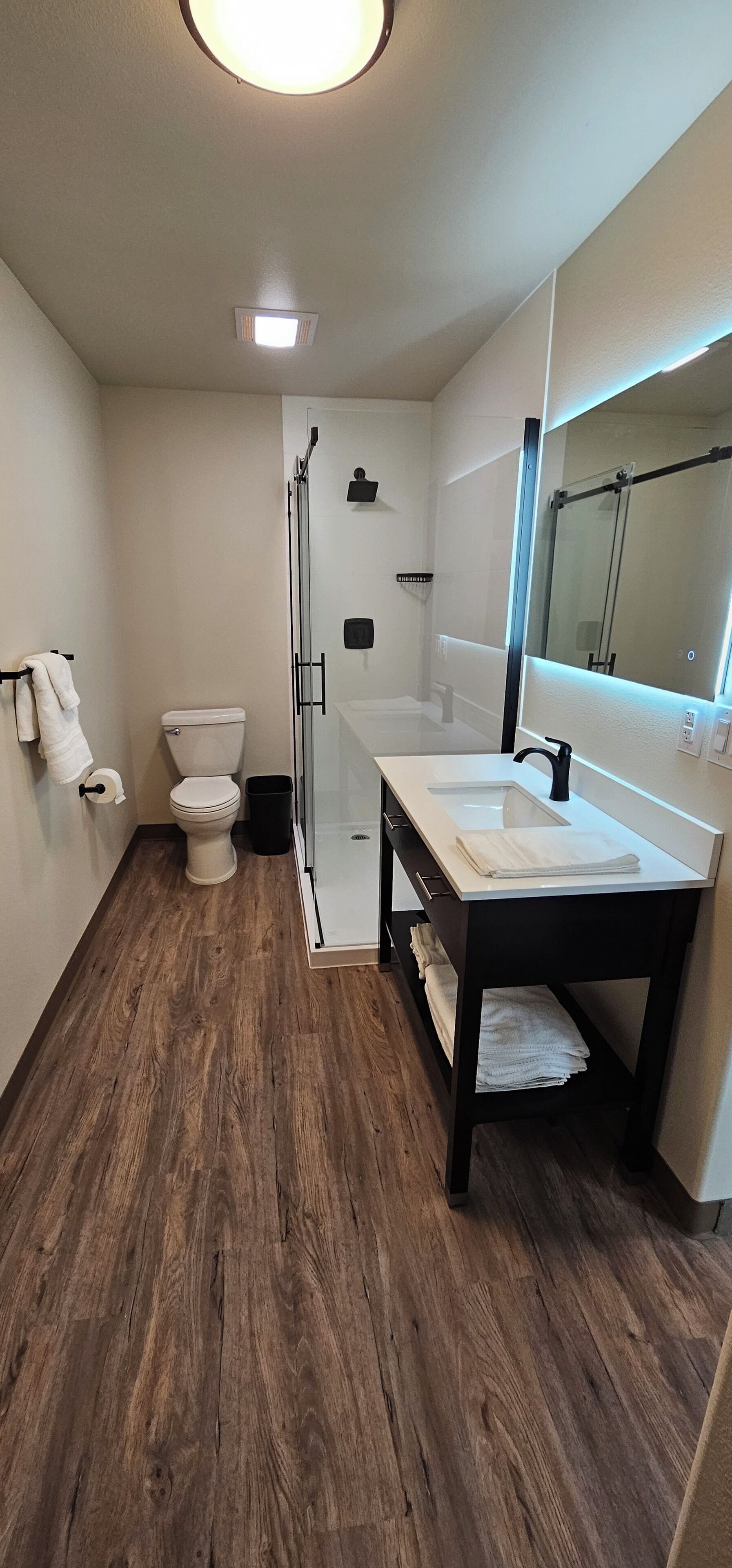 Un baño moderno con suelo de madera, inodoro blanco, cabina de ducha de vidrio y tocador con espejo.