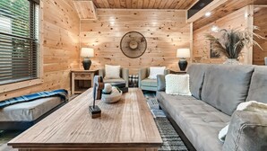 Un acogedor interior de cabaña con techo de madera, una mesa central con objetos decorativos, un sofá gris y una ventana con persianas.