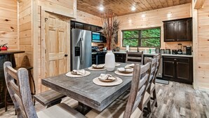 Cocina de cabaña de madera con una mesa de comedor dispuesta para seis personas, un refrigerador, un microondas y una ventana con vistas a la vegetación.