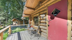Punaisella ovella varustettu hirsihökkelö, jossa on puinen terassi ulkokalusteineen ja pieni ikkuna, josta avautuu näkymä puihin.