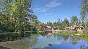 A serene lakeside setting with two wooden cabins, clear water reflecting the surrounding trees, and a dock with a kayak.
