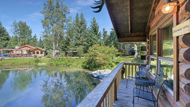 A cabin with a wooden deck overlooking a lake, a sign reading "Cottonwood Valley," and a boat docked nearby.