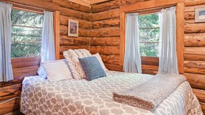 Ein Blockhausszimmer mit Bett, zwei mit Vorhängen versehenen Fenstern, einem gerahmten Bild an der Wand und einer Holzbalkendecke.