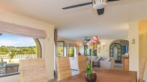 A dining area with a wooden table, wicker chairs, and a vase with flowers. There is a ceiling fan above and a view of the outdoors through large windows. - Villa Las Viñas with swimming pool & stunning view (Teulada)