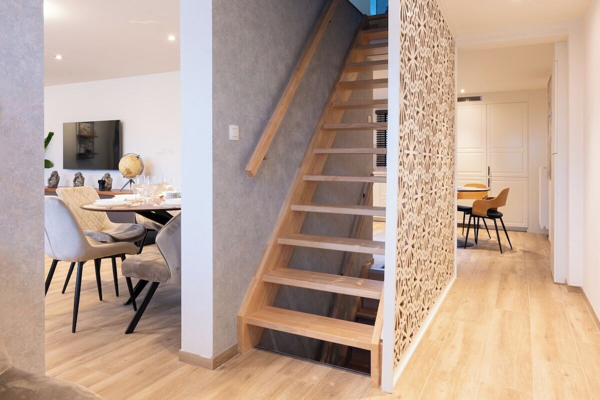 A modern interior with a wooden staircase, a dining area with chairs and a table, and a kitchen in the background.