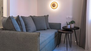 A modern, minimalist living room with a grey sofa, a small round table with a plant, and two wall-mounted lights. - Silo Luxury Rooms Nafplio (Argos - Mykines)