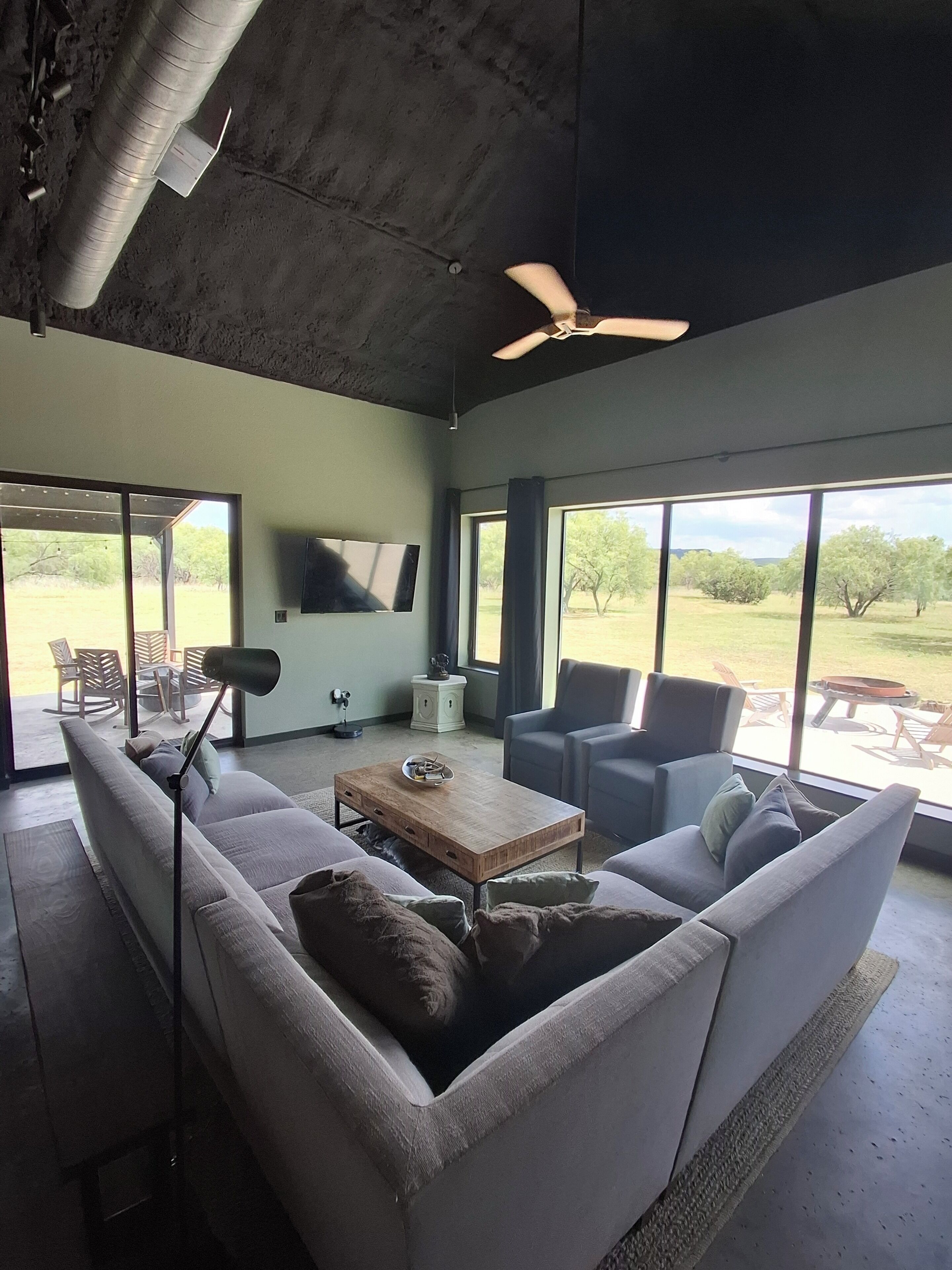 Una sala de estar moderna con sofá, sillones, una mesa de centro, televisor y ventanas grandes con vistas al exterior.