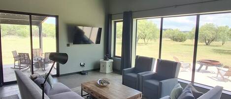 A modern living room with a sofa, chairs, a coffee table, a TV, and large windows with a view of the outdoors.