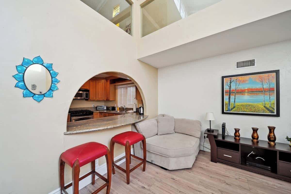A modern living room with a sofa, two red bar stools, a painting, a wooden cabinet, and a kitchen area with a window.