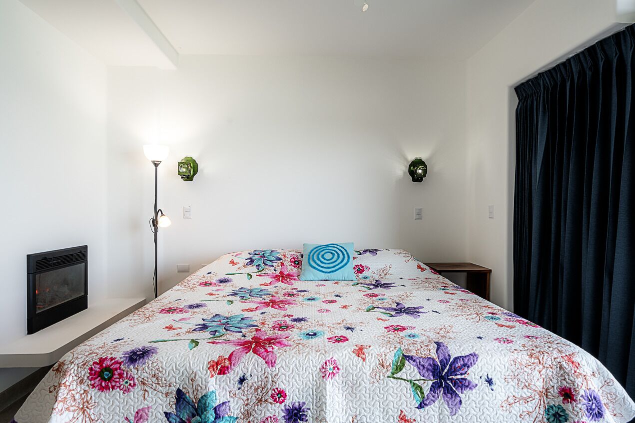 Un dormitorio con colcha floral, chimenea, dos lámparas de pared y una cortina oscura.