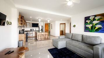 A modern living room with a grey sectional sofa, a wooden coffee table, and a dark blue rug. There is a kitchen area with wooden cabinets and a dining space with a wooden table and chairs.