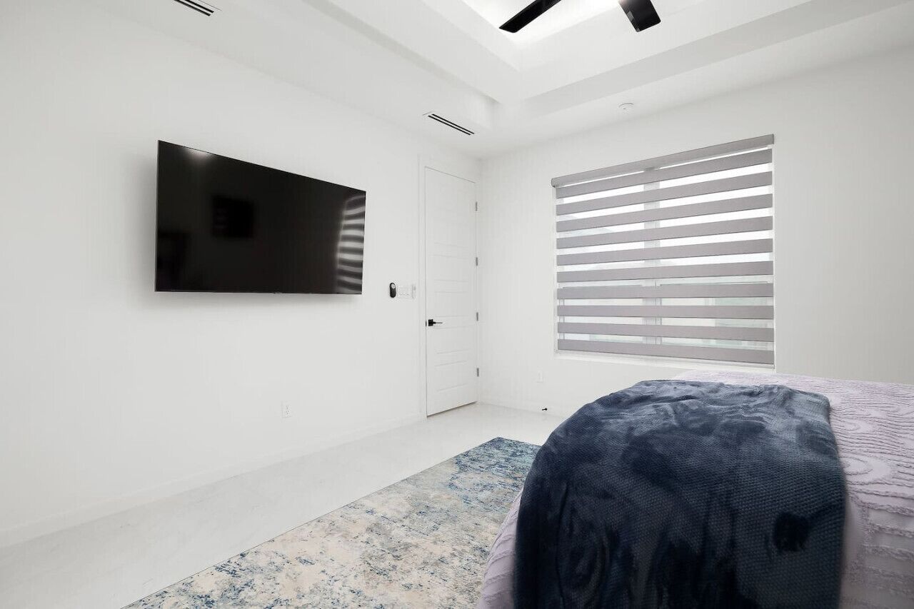 A modern bedroom with a flat-screen TV mounted on the wall, a large window with blinds, and a patterned rug on the floor.